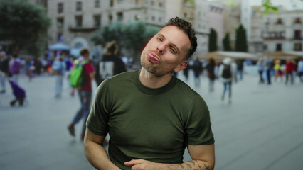 Young man with tattoos making funny faces in an urban square with people walking in the background,...