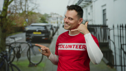 Young man wearing a red volunteer shirt smiles and gestures talking on phone while pointing on a...