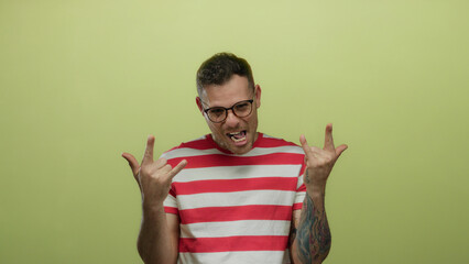 Young hispanic man in glasses making rock gesture on yellow background with tattooed arm and striped shirt exuding an energetic and playful vibe during isolated celebration