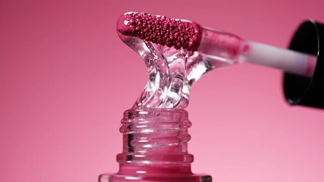 Close up of Clear Lip Gloss Dropping from Applicator into Bottle with Pink Background and Soft Lighting Showing Fine Details of Product Texture and Shine