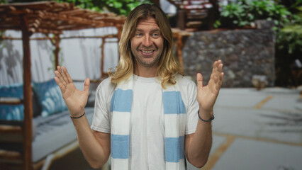 Blond man with long hair smiles and raises arms at restaurant on street wearing striped fan scarf; team pride.