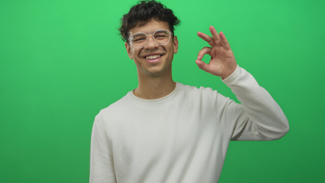 Man wearing glasses facing camera and smiling shows ok sign with hand in a bright green studio; approval. - Powered by Adobe