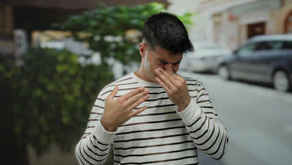 Young man in striped shirt covering nose while standing in a city street during the day.