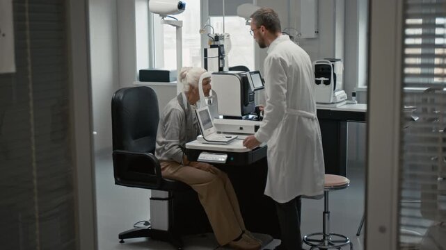 Side view shot of male ophthalmologist assisting senior woman during eye exam in clinic and setting up modern diagnostic machine, copy space