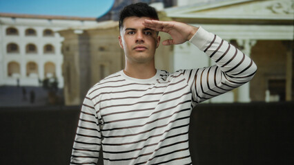 Young man saluting at university courtyard with historic architecture wearing striped shirt on a sunny day