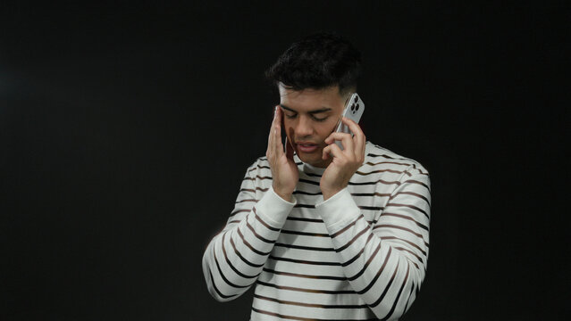 Young man in striped sweater talks on smartphone with concerned expression against black background.
