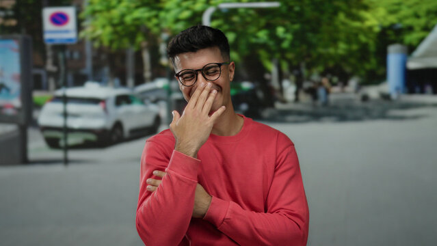 Young man wearing glasses in a red sweater smiles joyfully at the camera on a sunny city street with trees and cars in the background. - Powered by Adobe