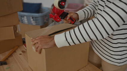 Man packing cardboard box in living room of new home with tape dispenser amid unpacked household items.