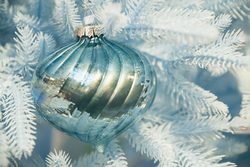 Teal glossy bauble on pastel blue Christmas tree with warm lighting. Festive decoration closeup in soft winter palette.
