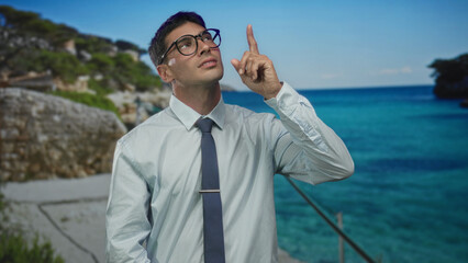 Young man pointing finger at sky beside rocky seaside path with turquoise sea and stone wall under clear sky; hope.