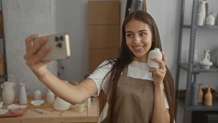Young woman holding ceramic vase and smartphone with bare hand, fingers tapping while smiling in pottery studio; creative joy.