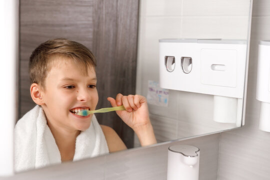 A young boy with a towel over his shoulder smiles while brushing his teeth in front of a modern bathroom mirror, focusing on good hygiene and daily routine.