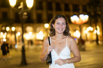 Obraz premium Long-haired cheerful female brunette walks along city street, spends time in motion, walks and enjoys sunny warm day, walks from university to home. Carefree, night cafe, promenade.