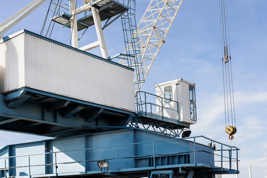 Industrial crane structure rises under clear sky, showcasing metal framework, cabin, pulley and hook. Scene highlights machinery, steel geometry and port atmosphere with strong sunlight and shadows