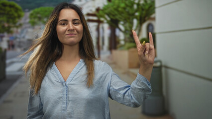 Fototapeta premium Woman smiling with right hand showing rock horns gesture on a city street beside planter and trashcan near storefront; joy fun casual.