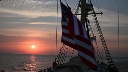 American flag flutters in the wind on a sailing ship, silhouetted against a beautiful sunset over the sea. The image evokes feelings of freedom, adventure, and patriotism Stock Video - Powered by Adobe