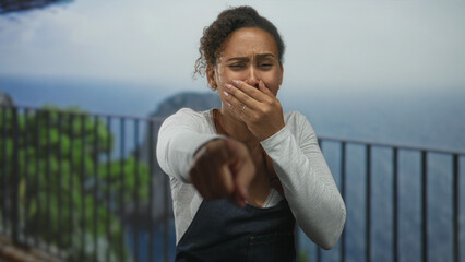 Fototapeta premium Woman covering mouth with hand and pointing finger toward camera at seaside railing; shock reaction.