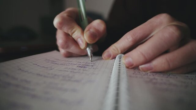 A person writes down notes in a spiral notebook using a pen while sitting in a cozy, softly lit space in the evening hours.