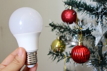 Hand holding LED light bulb with decorated Christmas tree in background. Electricity bill concept during the Christmas holiday. 