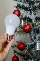 Hand holding LED light bulb with decorated Christmas tree in background. Electricity bill concept during the Christmas holiday. 