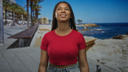 Fototapeta premium Woman with exposed midriff tilts head back on street promenade facing ocean and palm lined boardwalk, eyes closed and smiling in warm sunlight; serenity.