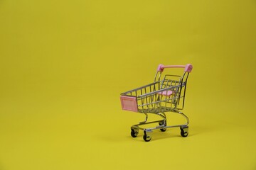 A small pink shopping cart sits against a yellow backdrop. Ideal for concepts of shopping, e-commerce, gift ideas, promotions, and family-friendly branding.
