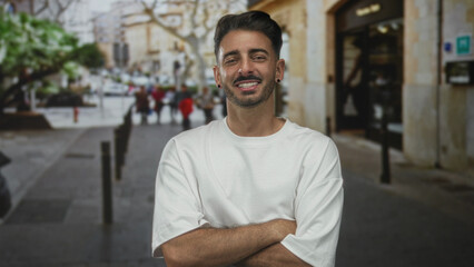Hispanic man in white t shirt stands with bare arms crossed on busy sunlit city street; confidence...