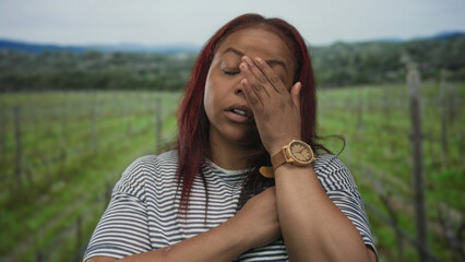 Obraz premium Woman resting head on hand with eyes closed in forest among vineyard stakes wearing striped shirt; exhaustion.