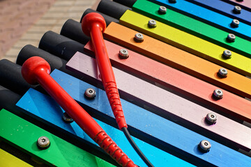 A vibrant close-up of a colorful outdoor xylophone with two bright red mallets resting on the rainbow-colored keys in a public park, emphasizing musical play and childhood fun.
