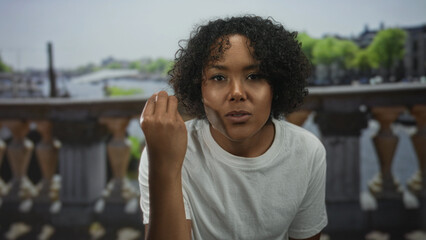 Woman holds clenched fist by canal outdoors in urban street scene while looking ahead;...