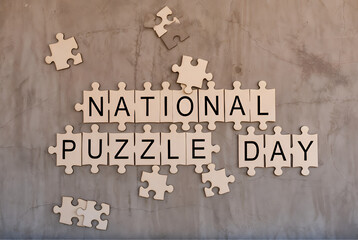 Wooden jigsaw puzzle pieces spelling National Puzzle Day arranged on textured concrete wall.