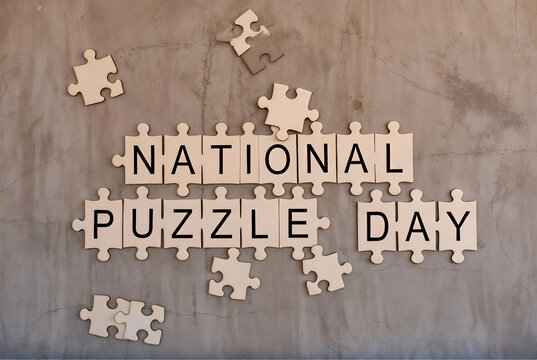 Wooden jigsaw puzzle pieces spelling National Puzzle Day arranged on textured concrete wall.
