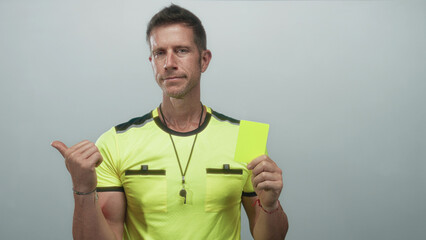 Man referee in neon yellow shirt holding a bright yellow card while thumb points to card in light gray studio; caution.