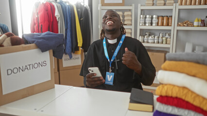 Man holding smartphone by a donations box in a building, fist pump and surprised face while seated at donation center; community service joy.