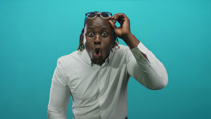 Man lifting glasses in teal studio with open mouth and surprised eyes; surprise reaction response...