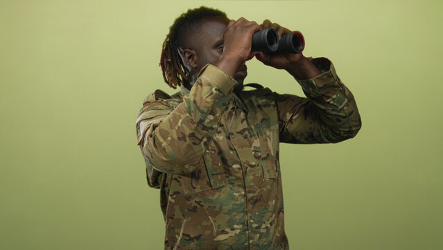 Man in soldier uniform holds binoculars to eyes against a pale green studio backdrop, wearing camouflage jacket and dog tag; vigilance duty. - Powered by Adobe