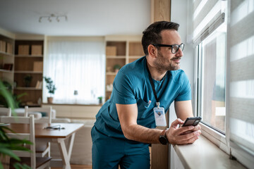 Male healthcare professional relaxing looking out window holding smartphone