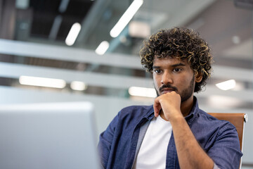 Close-up photo of a young Indian man sitting thoughtfully at an office desk and looking at a laptop screen