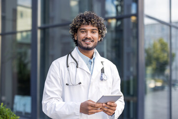 Portrait of a young smiling Indian male doctor in a white coat standing outside, holding a tablet and looking at the camera