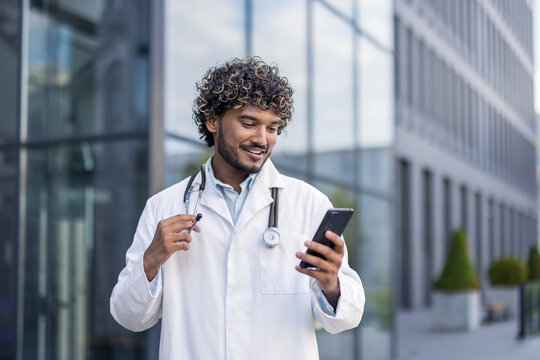 Young Indian male doctor in white coat standing outside clinic and using mobile phone - Powered by Adobe