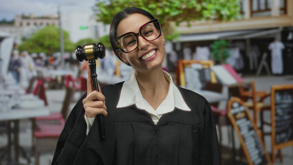 Hispanic woman judge holding gavel smiling playfully on restaurant terrace wearing uniform with...