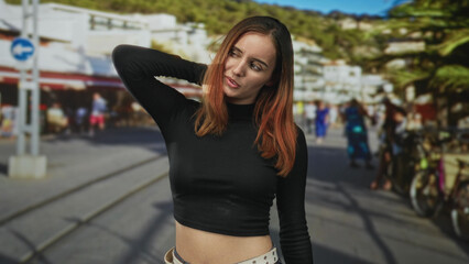 Young redhead woman with hand behind head showing midriff, wearing black croptop, standing by bicycles and cafe terraces on a busy street; pensive contemplation.
