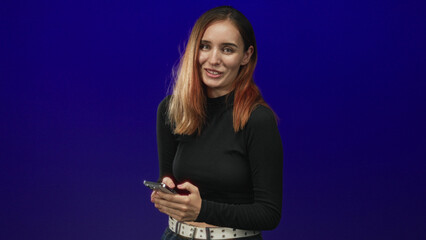 Young redhead woman holding smartphone and texting with hands, smiling in blue studio wearing black long sleeve top and white belt; friendliness.
