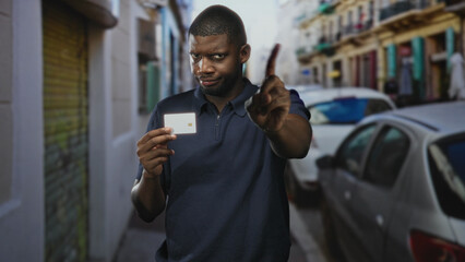 Young black man holds creditcard and points finger at camera in narrow street with parked cars and...