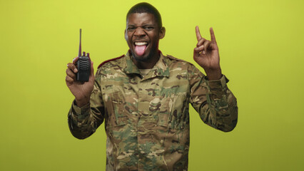 Soldier in camouflage uniform holding walkie talkie and making horns hand gesture while smiling in...
