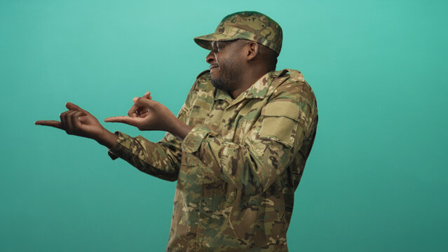 Man soldier in camouflage uniform and cap points both index fingers outward while smiling in studio; confidence. - Powered by Adobe