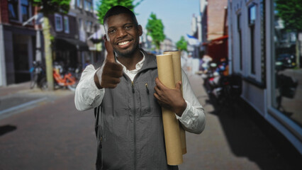 Man holding rolled blueprints and giving thumbs up gesture on a street in a city with storefronts and bicycles; confidence success.
