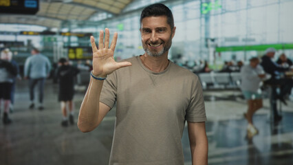 Man points index finger up in airport terminal showing hand and forearm while smiling to camera;...