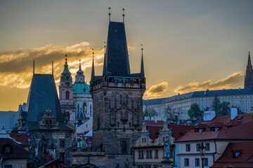 Fototapeta premium Sunset view of historic Prague with the Charles Bridge tower, old rooftops, and church domes glowing in warm evening light.