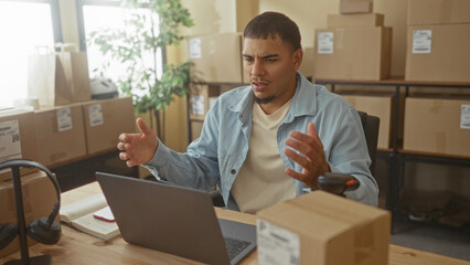 Man at laptop gesturing with hands amid stacked boxes, barcode scanner and headset in packaging...